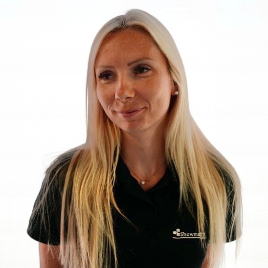 Eine Frau mit langen blonden Haaren trägt ein schwarzes Poloshirt mit einem Logo darauf. Sie blickt leicht nach links vor einem strahlend weißen Hintergrund.