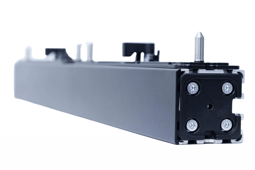 Nahaufnahme eines schwarzen, rechteckigen mechanischen Bauteils mit vier sichtbaren Schrauben an einem Ende, geringe Tiefenschärfe. Es weist verschiedene Vorsprünge und einen Metalldorn an der Oberseite auf, vor einem schlichten weißen Hintergrund.