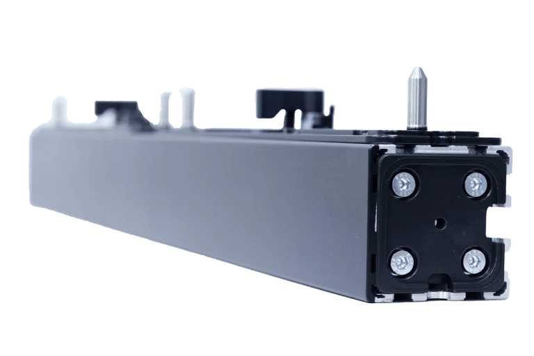 Nahaufnahme eines schwarzen, rechteckigen mechanischen Bauteils mit vier sichtbaren Schrauben an einem Ende, geringe Tiefenschärfe. Es weist verschiedene Vorsprünge und einen Metalldorn an der Oberseite auf, vor einem schlichten weißen Hintergrund.