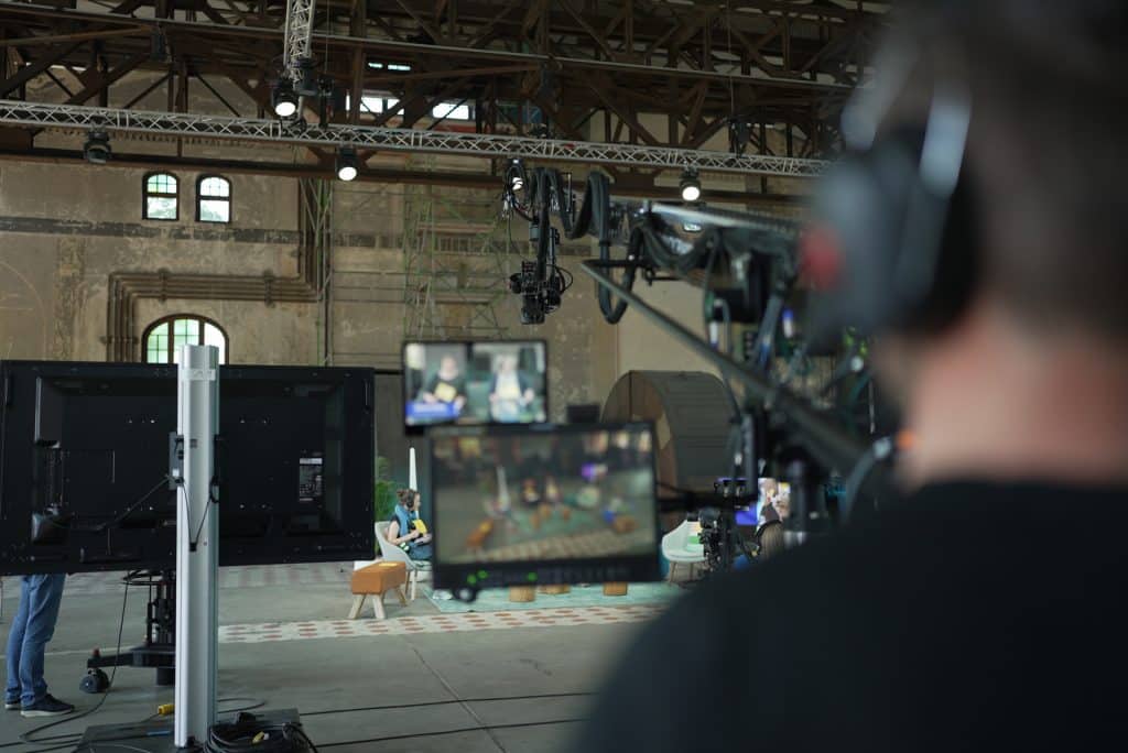 Ein Blick hinter die Kulissen eines Videoproduktionssets mit Kameras und Crew. Im Vordergrund trägt eine Person Kopfhörer, während im Hintergrund eine andere Person auf einem orangefarbenen Stuhl in einem Raum im Industriestil sitzt.