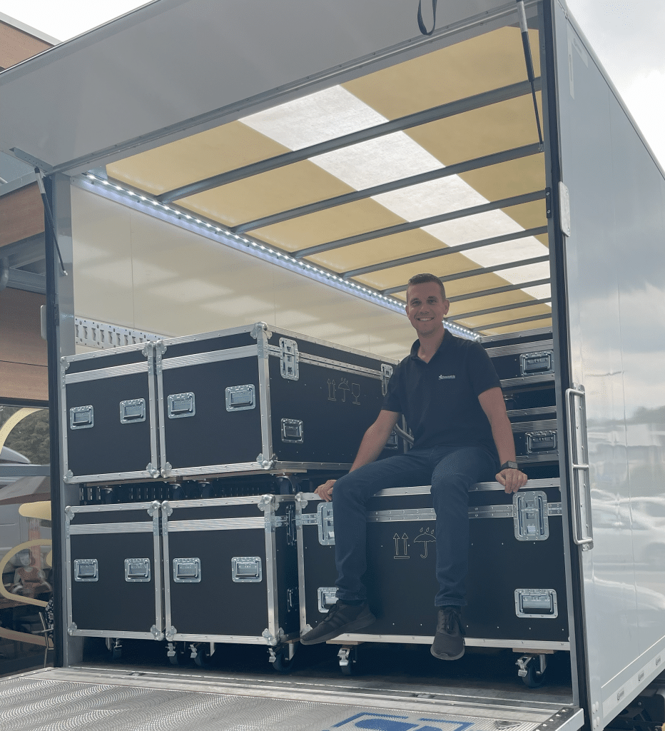 Eine Person in einem schwarzen Hemd sitzt auf der Kante eines offenen Lastwagens, der mit mehreren gestapelten schwarzen Ausrüstungskoffern beladen ist. Der Lastwagen hat ein transparentes Dach, das Licht ins Innere lässt. Die Szene scheint im Freien zu sein.