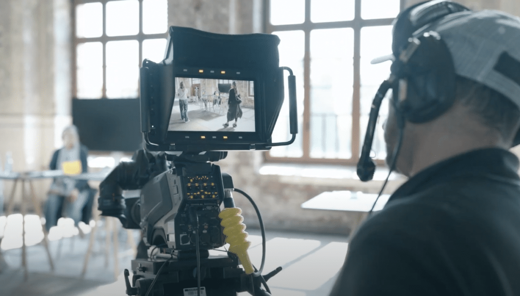 Eine Person bedient in einem Innenraum eine professionelle Videokamera, die auf einem Stativ montiert ist. Sie trägt ein Headset und eine Kappe und konzentriert sich auf den Kamerabildschirm, auf dem Personen in einem Raum mit großen Fenstern im Hintergrund zu sehen sind.