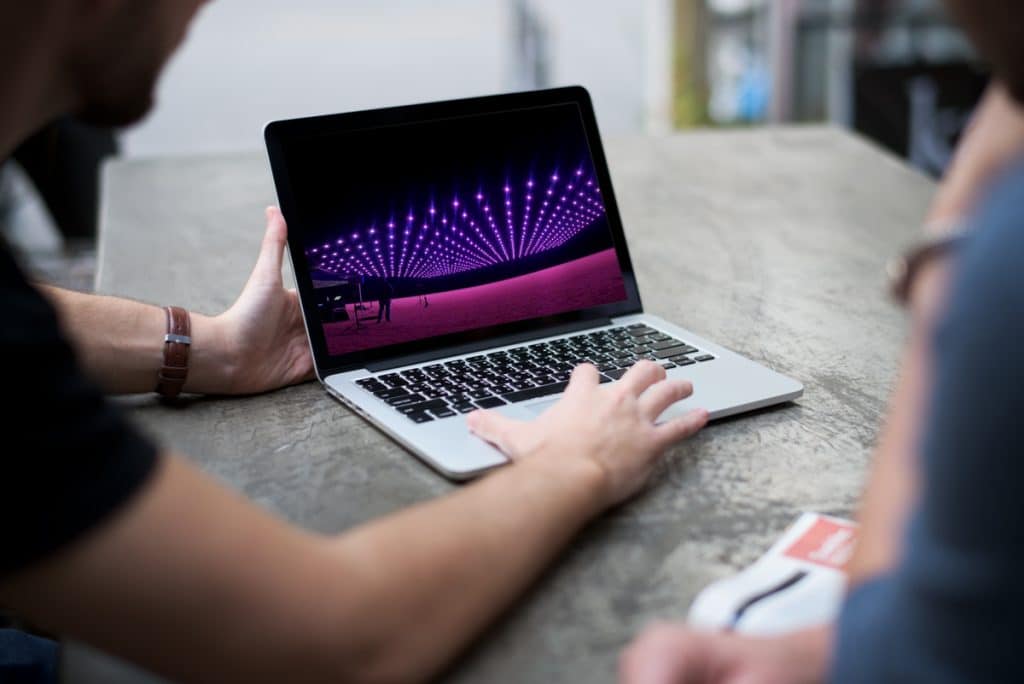 Zwei Personen sitzen an einem Tisch, eine davon benutzt einen Laptop. Auf dem Laptop-Bildschirm ist ein lebendiges Bild einer futuristischen Szene mit violetten Lichtern zu sehen, die in einem Muster angeordnet sind und an eine moderne digitale Installation oder Lichtshow erinnern.