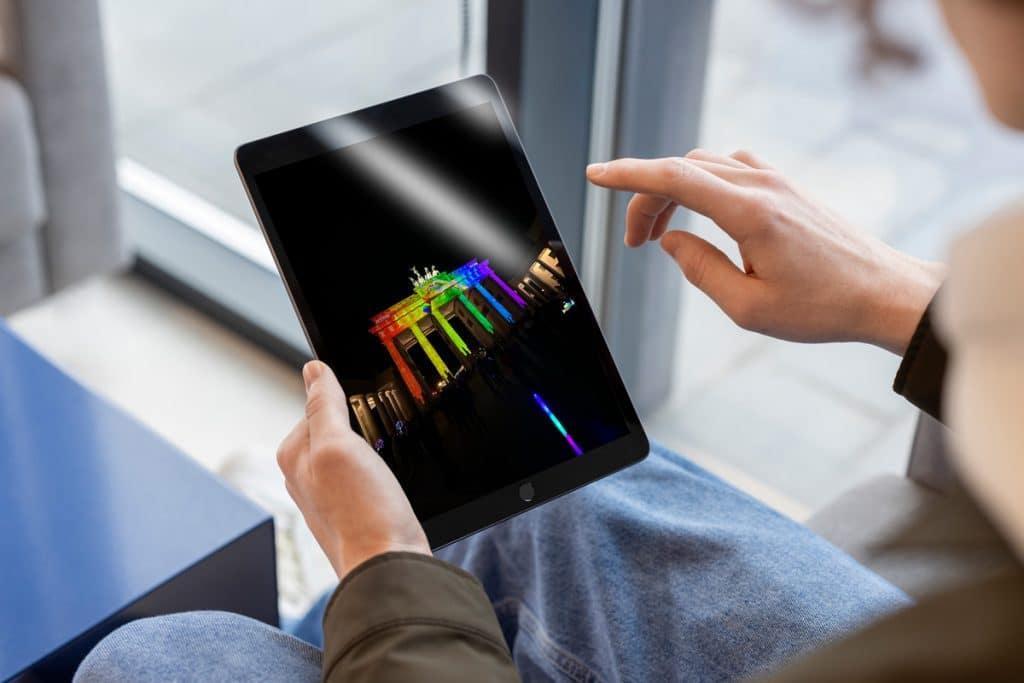 Person hält ein Tablet, auf dem das Bild eines Denkmals in Regenbogenfarben angezeigt wird. Die Person sitzt im Innenbereich neben einem großen Fenster und trägt eine braune Jacke und hellblaue Jeans.