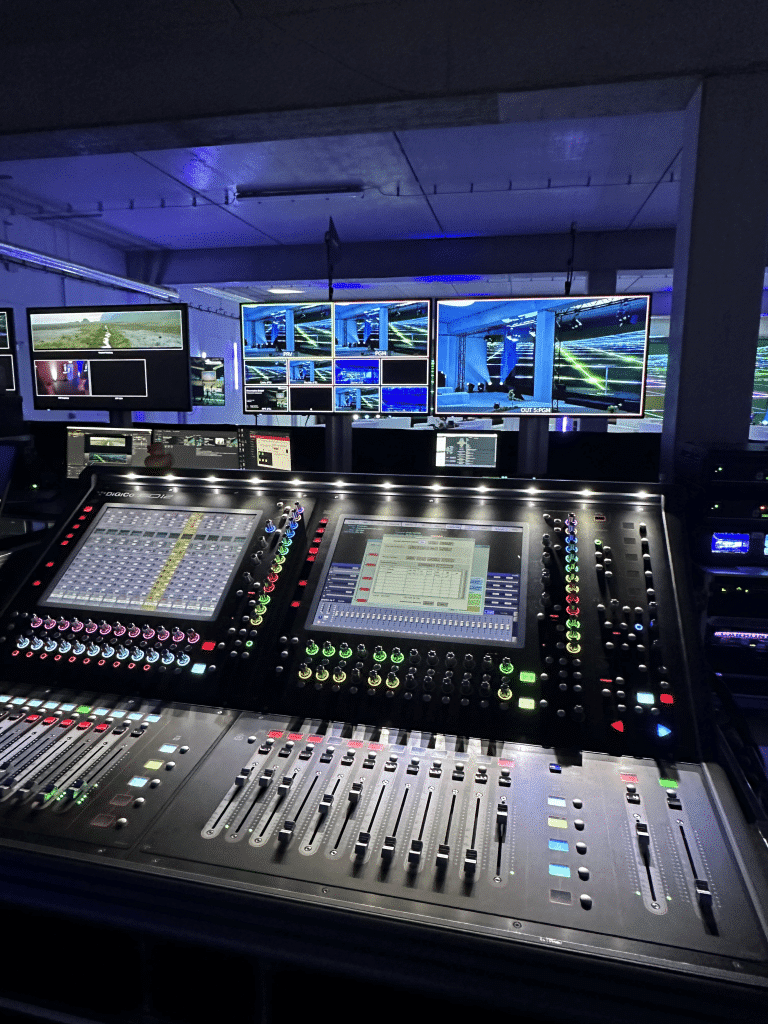 Ein schwach beleuchteter Kontrollraum mit einem Audiomischpult mit zahlreichen Knöpfen, Schiebereglern und digitalen Bildschirmen. Mehrere Monitore zeigen im Hintergrund verschiedene Video-Feeds eines Live-Events oder einer Live-Übertragung an.